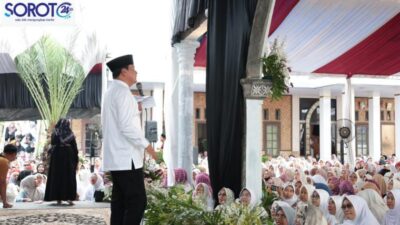 Seorang pejabat pria memakai baju putih dan peci hitam berdiri di panggung berbicara di depan ratusan jamaah perempuan berhijab