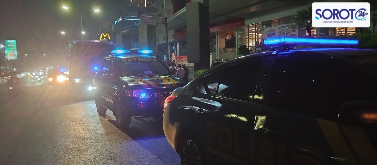 Dua mobil polisi warna hitam dengan lampu biru menyala melintas di jalan raya malam hari di depan pusat perbelanjaan
